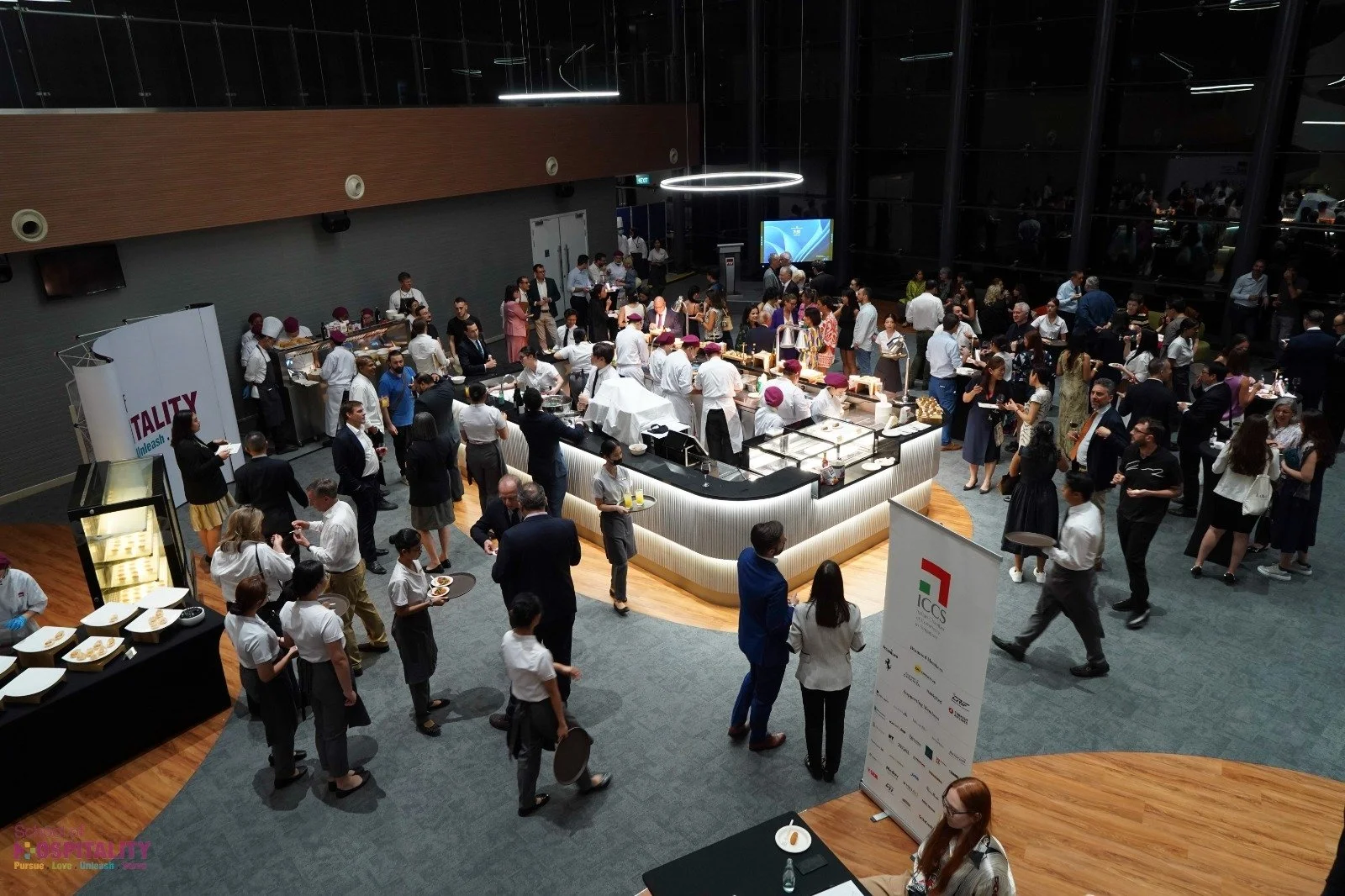 A large indoor event with a crowd of people at a buffet-style food station. Attendees are socializing and serving themselves food, with some waiting in line. The venue has modern lighting, wooden and carpeted flooring, and large glass windows.