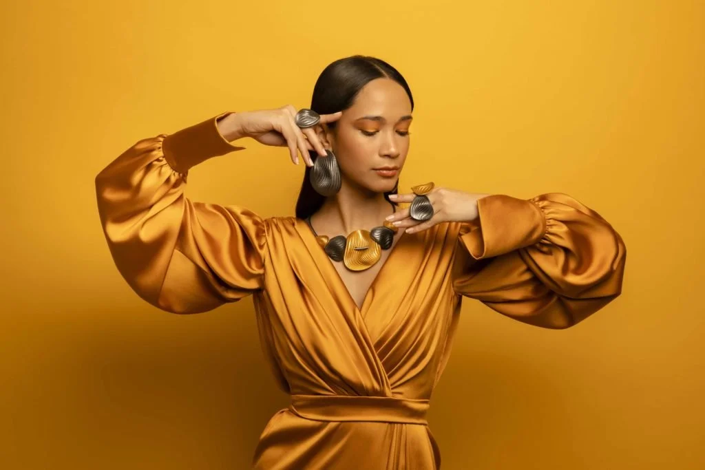 Woman in a shiny gold dress with matching jewelry, posing against a yellow background.