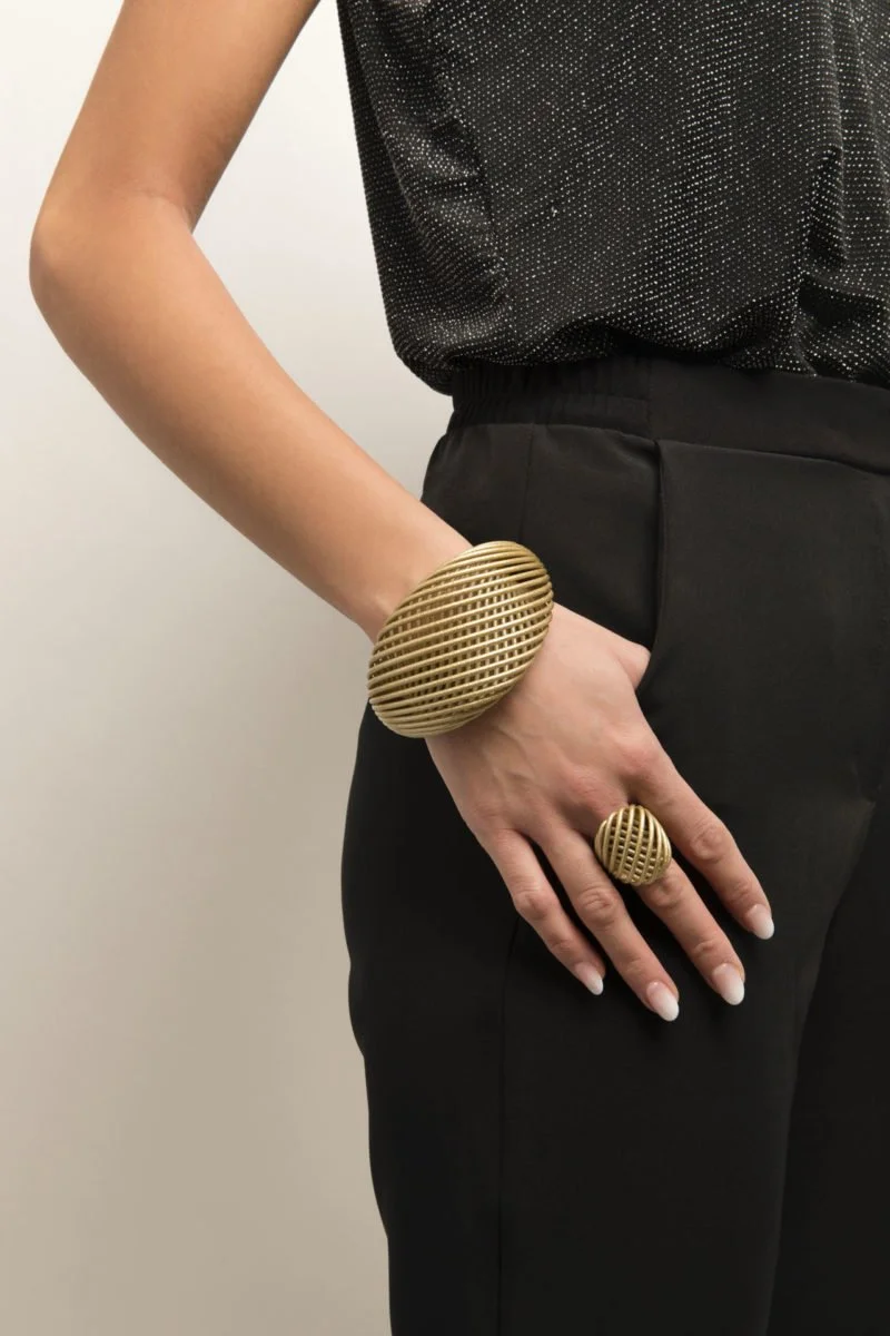 Person wearing black clothing, accessorized with large gold bracelet and ring with a grid pattern.