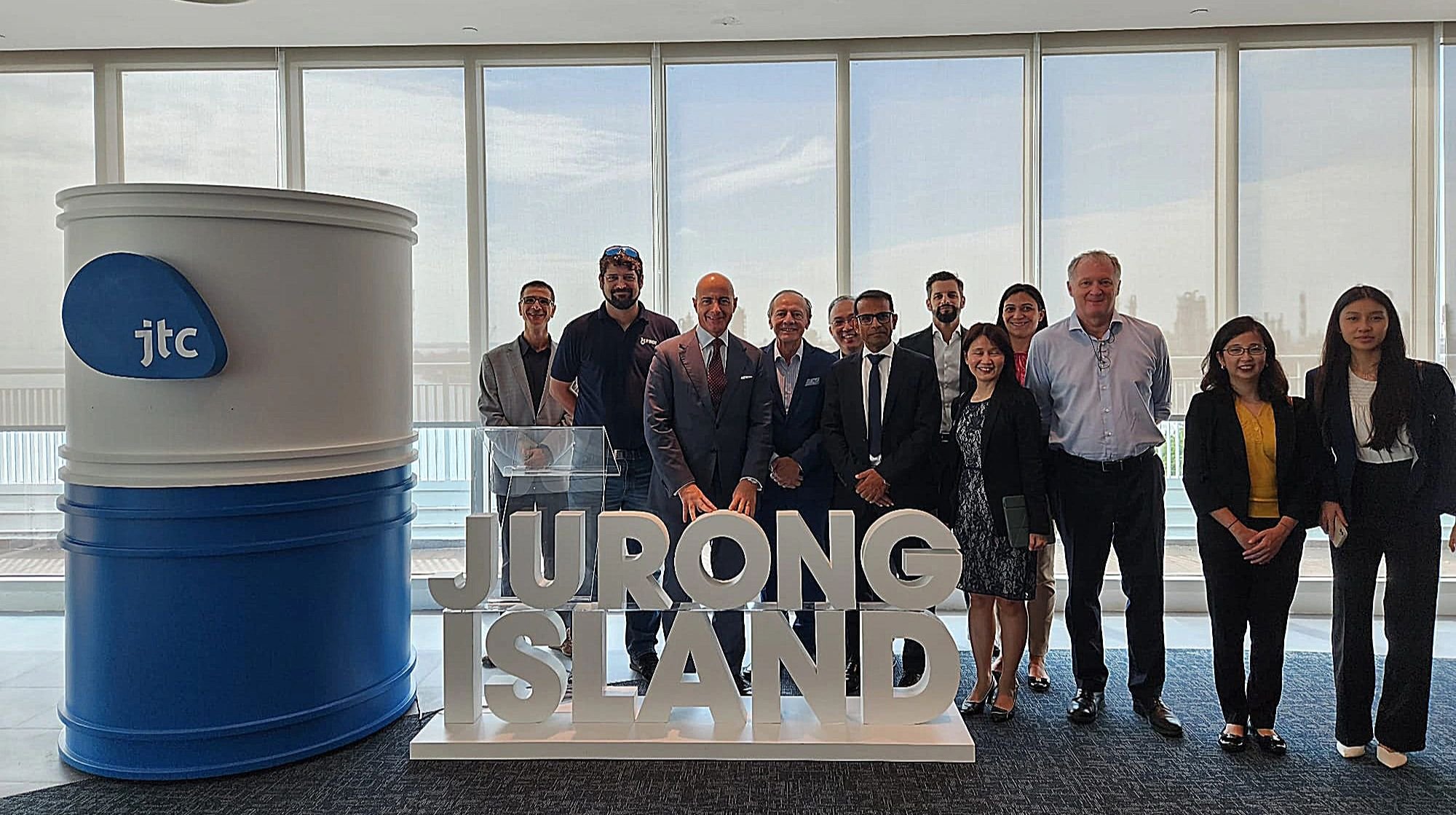 Group of people standing behind a sign that reads 'JURONG ISLAND' in front of large windows, with a large blue and white drum with the JTC logo to the left.
