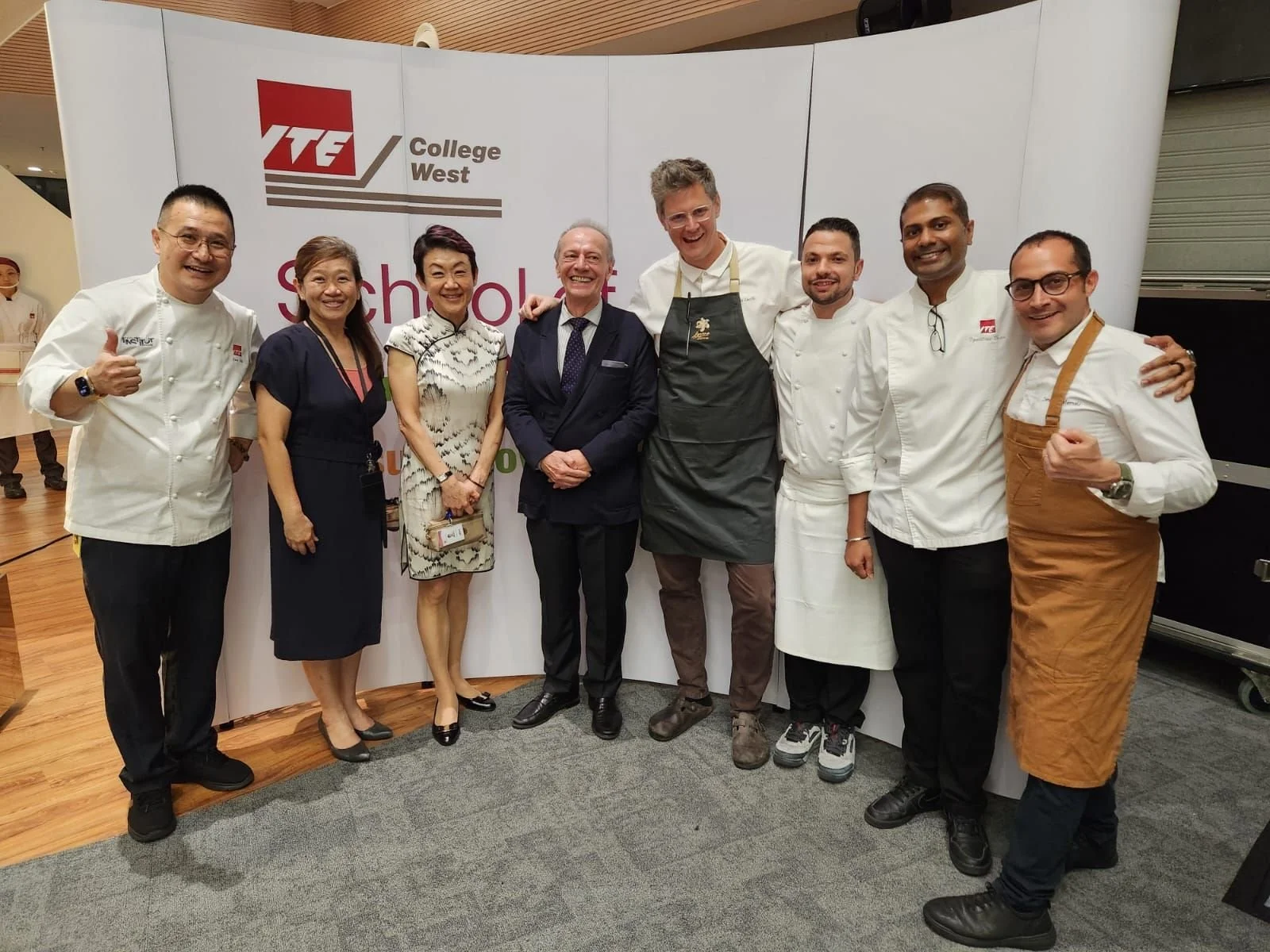 Group of nine people, chefs and officials, standing together in front of a white backdrop with the ITE College West logo, smiling and posing for the camera.