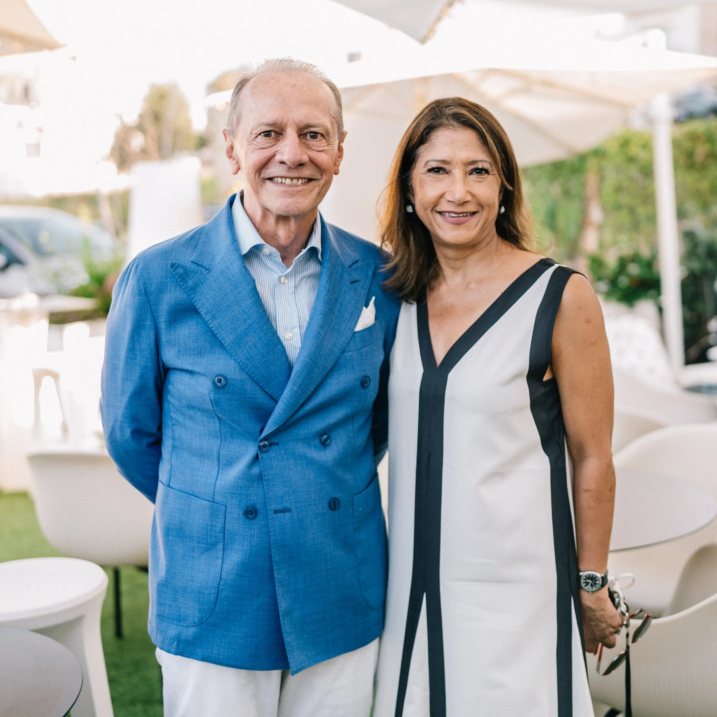 A smiling older man in a blue suit standing next to a smiling woman in a black and white sleeveless dress outdoors, with white chairs and umbrellas in the background.