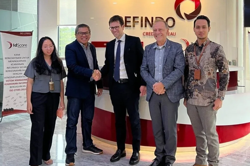 Five people in business attire standing in front of a reception desk with the logo of Pefindo Credit Bureau. Two of them are shaking hands, and there is a banner on the left side of the image.