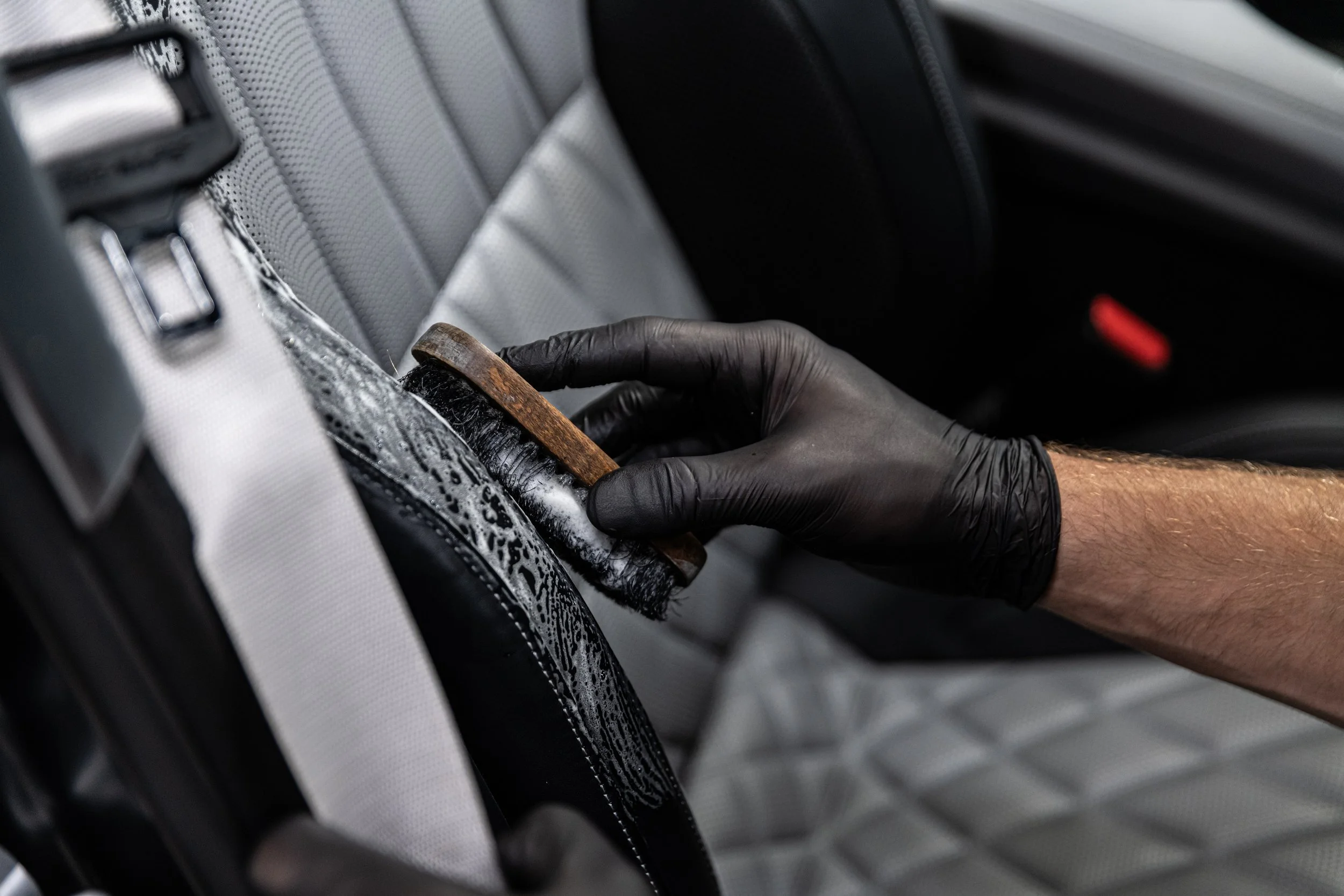 A person wearing a black glove is cleaning a car seat with a cloth and brush.
