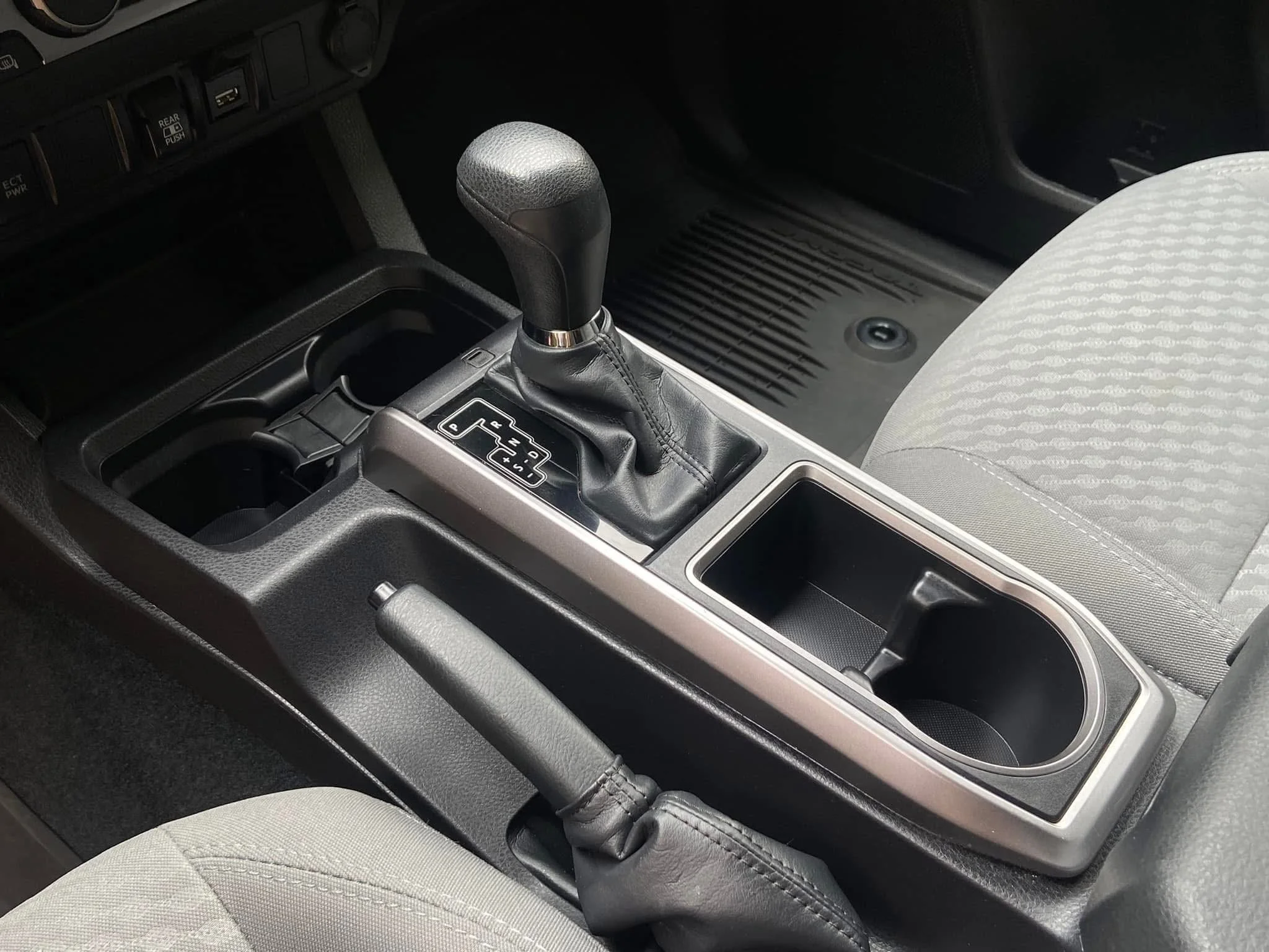 The image shows the center console of a vehicle, featuring the gear shift lever, two cup holders, and a small storage compartment. Part of the parking brake handle and the driver’s seat are also visible.