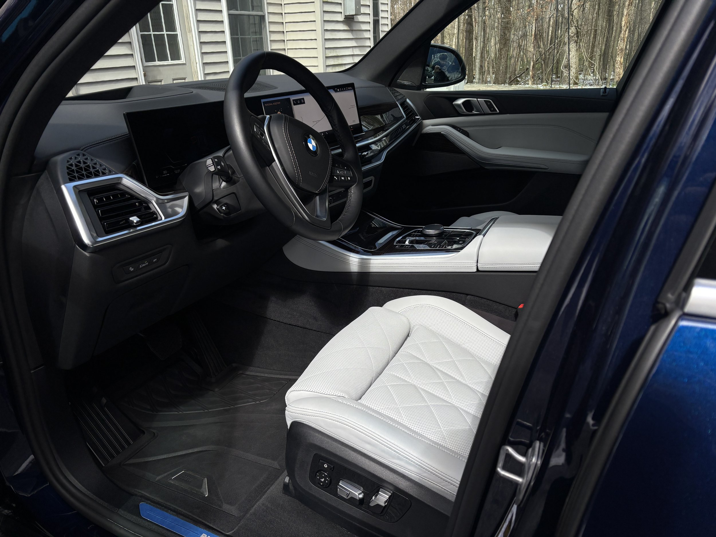 Interior of a luxury BMW car with a black dashboard, leather steering wheel, digital displays, and a white leather front passenger seat, parked outdoors near a house and wooded area.