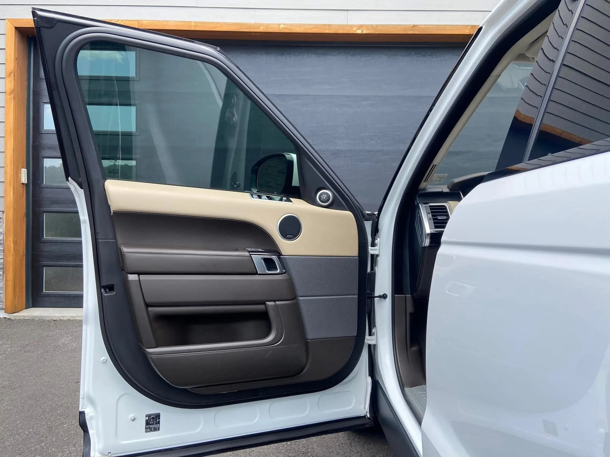 Open driver side door of a white vehicle showing beige and black interior door panel with a speaker and door handle, parked in front of a building with a wooden and gray facade.