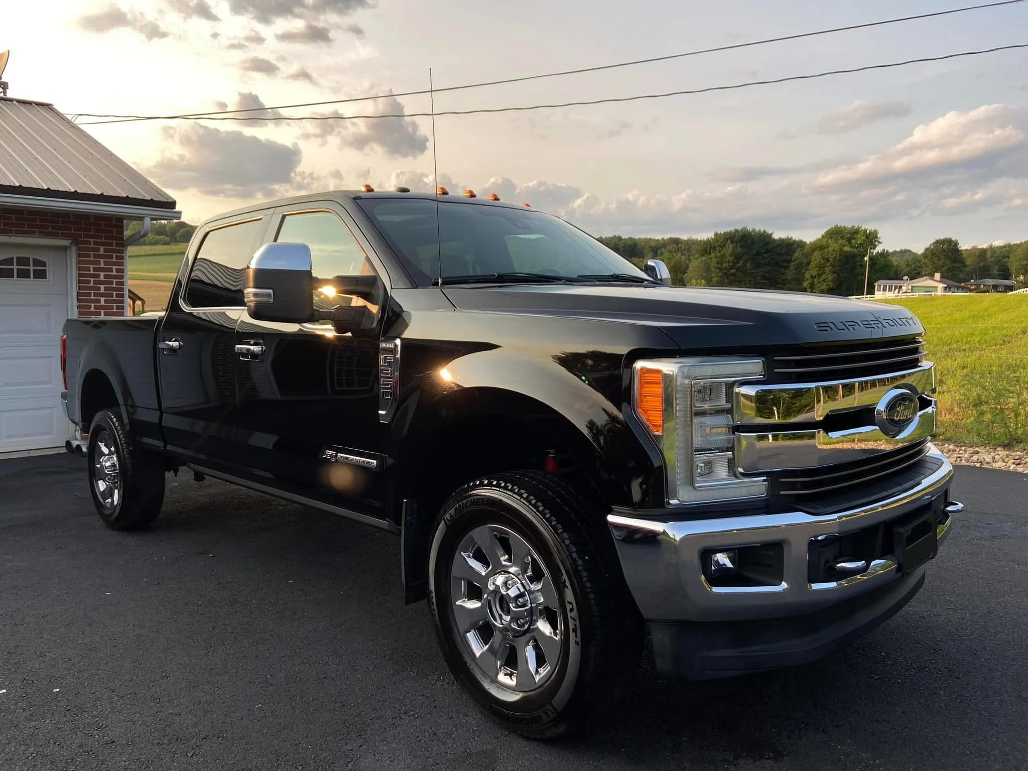 Black Ford Super Duty pickup truck parked on a driveway with a rural background and a garage to the left during sunset.