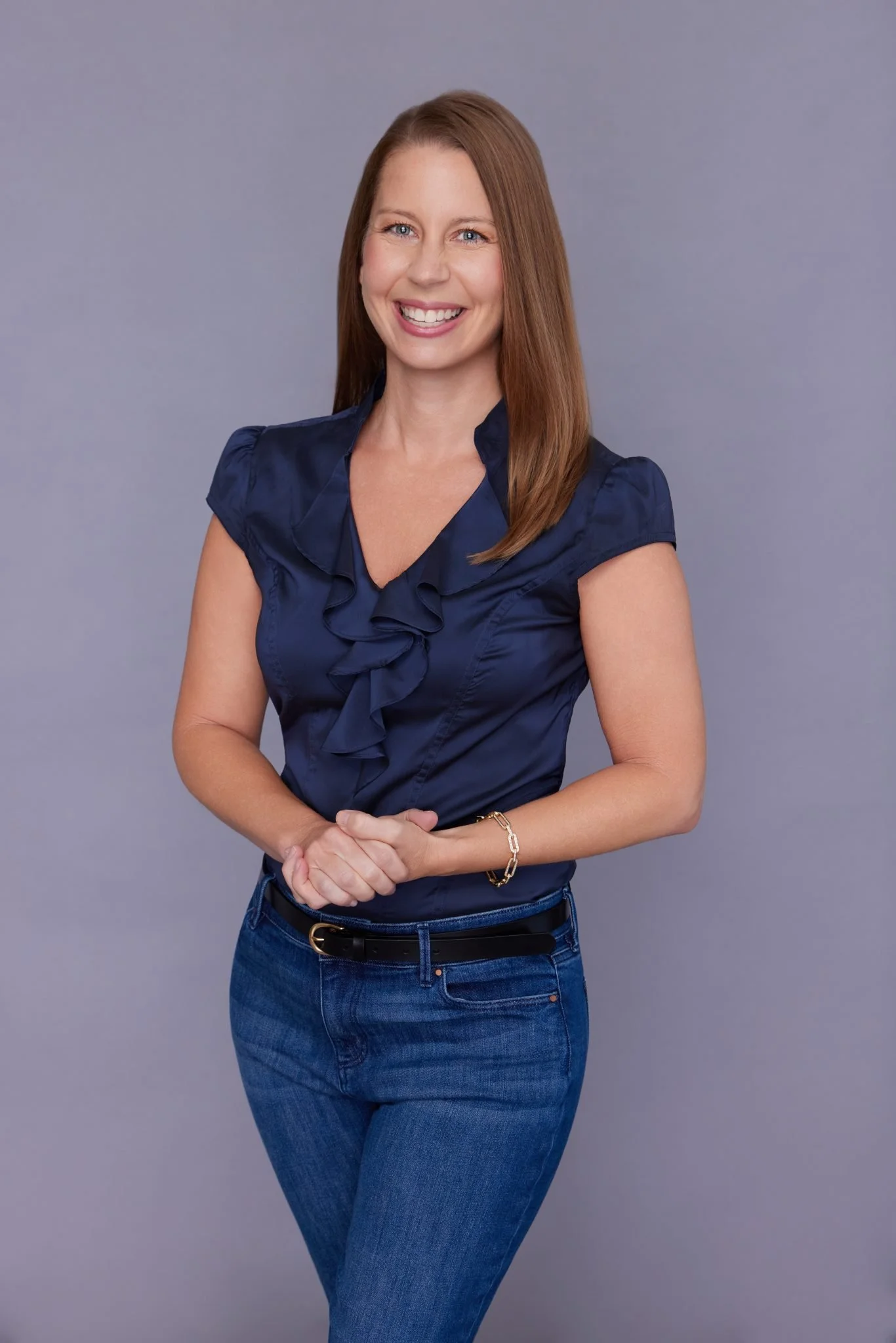 A woman with long brown hair, wearing a navy blue ruffled blouse and blue jeans, standing against a gray background, smiling with hands clasped.