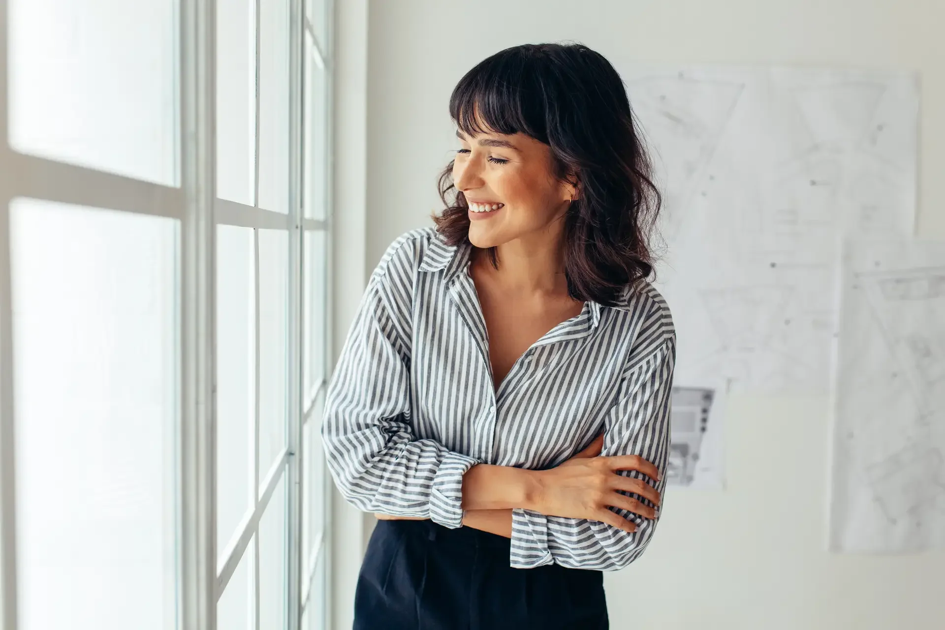 A woman with dark, wavy hair and a striped button-up shirt smiling as she looks out a large window in a bright, modern office or conference room.