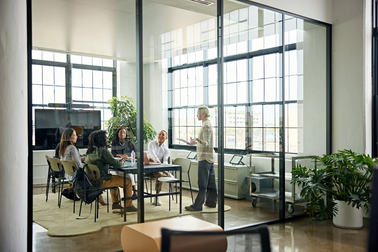 Business meeting in a glass-walled conference room with five women seated around a table and one standing woman presenting.