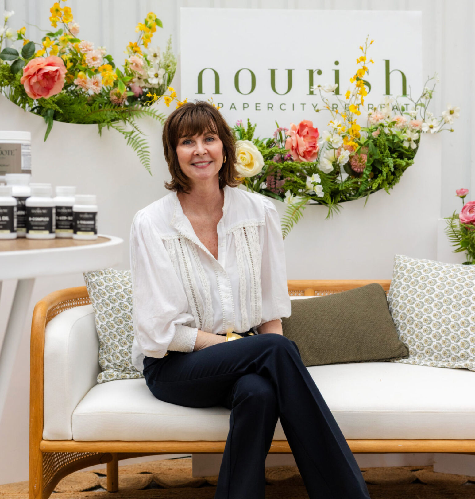 A woman with shoulder-length brown hair, wearing a white blouse and dark pants, is sitting on a cream-colored sofa with a wooden frame. Behind her is a large floral arrangement and a sign that reads 'nourish, PAPER CITY,' The woman is smiling and looking at the camera.
