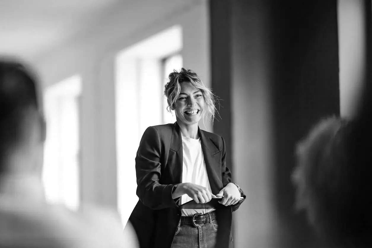 A woman smiling and speaking to a small group in an indoor setting with large windows, black blazer, white shirt, and messy short hair.