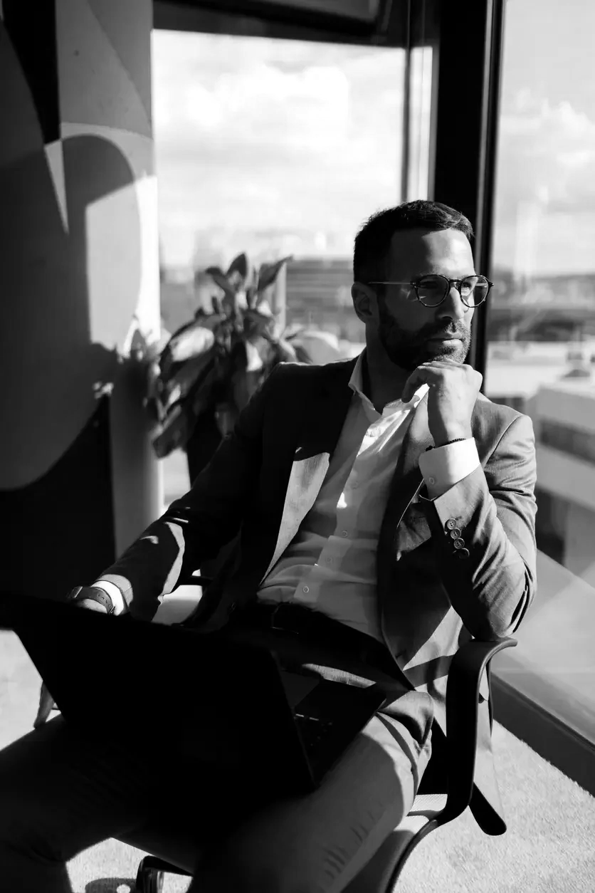 A man in professional attire sitting in an office chair near a large window, looking thoughtfully outside with a laptop on his lap, sunlight casting shadows in the room.