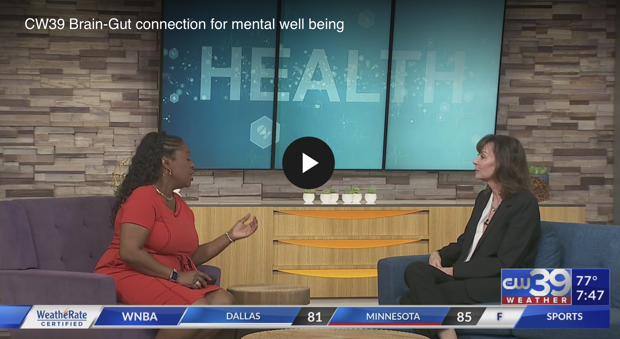 TV news interview with two women seated in a studio featuring a blue digital background with graphics and the word "HEALTH" prominently displayed. One woman is wearing a red dress, and the other is in a black blazer. The studio has a wooden panel with small potted plants on top and a weather update ticker at the bottom of the screen.