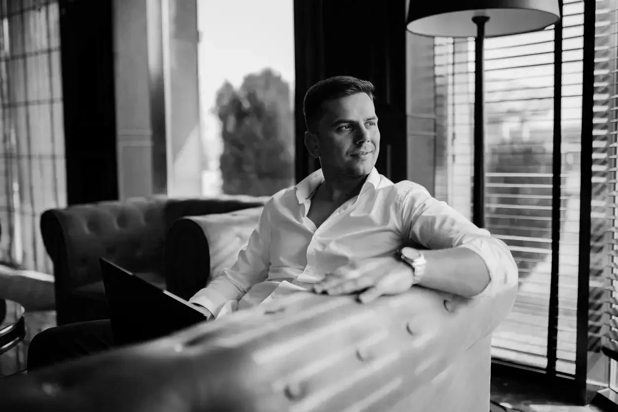 A young man in a white shirt sitting on a sofa, looking out a window.
