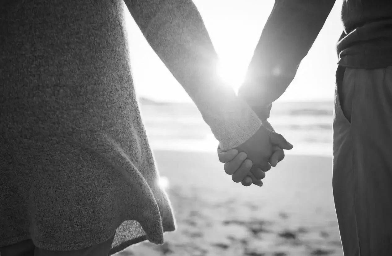 A black and white photo of two people holding hands on a beach at sunset, with the sun shining brightly behind them.