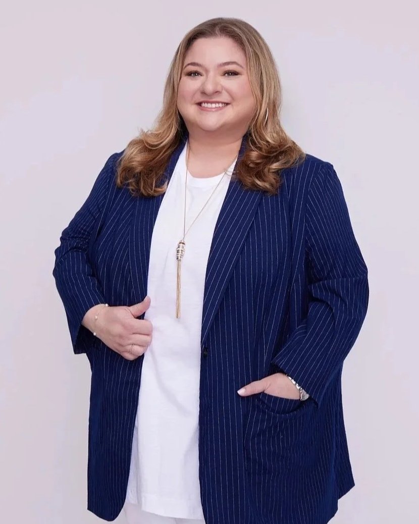 Woman with blonde hair, wearing a navy pinstripe blazer over a white shirt, smiling with one hand in her pocket and the other touching her blazer, against a plain light background.