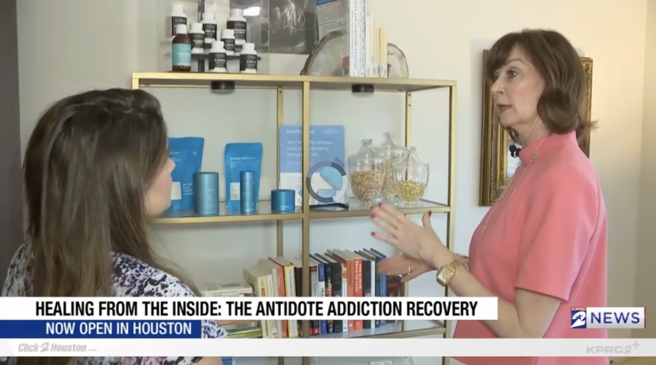 A woman in a pink blazer talking to another woman with long brown hair, who is wearing a patterned top, in front of a wooden bookshelf with bottles, jars, and books. A news banner at the bottom reads 'Healing from the Inside: The Antidote Addiction Recovery' and 'Now Open in Houston'.