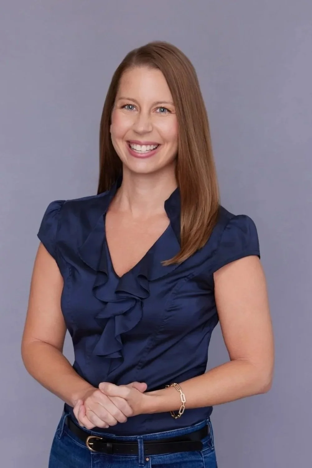 A woman with long, straight, light brown hair smiling, wearing a navy blue blouse with ruffles and blue jeans, standing against a plain gray background.