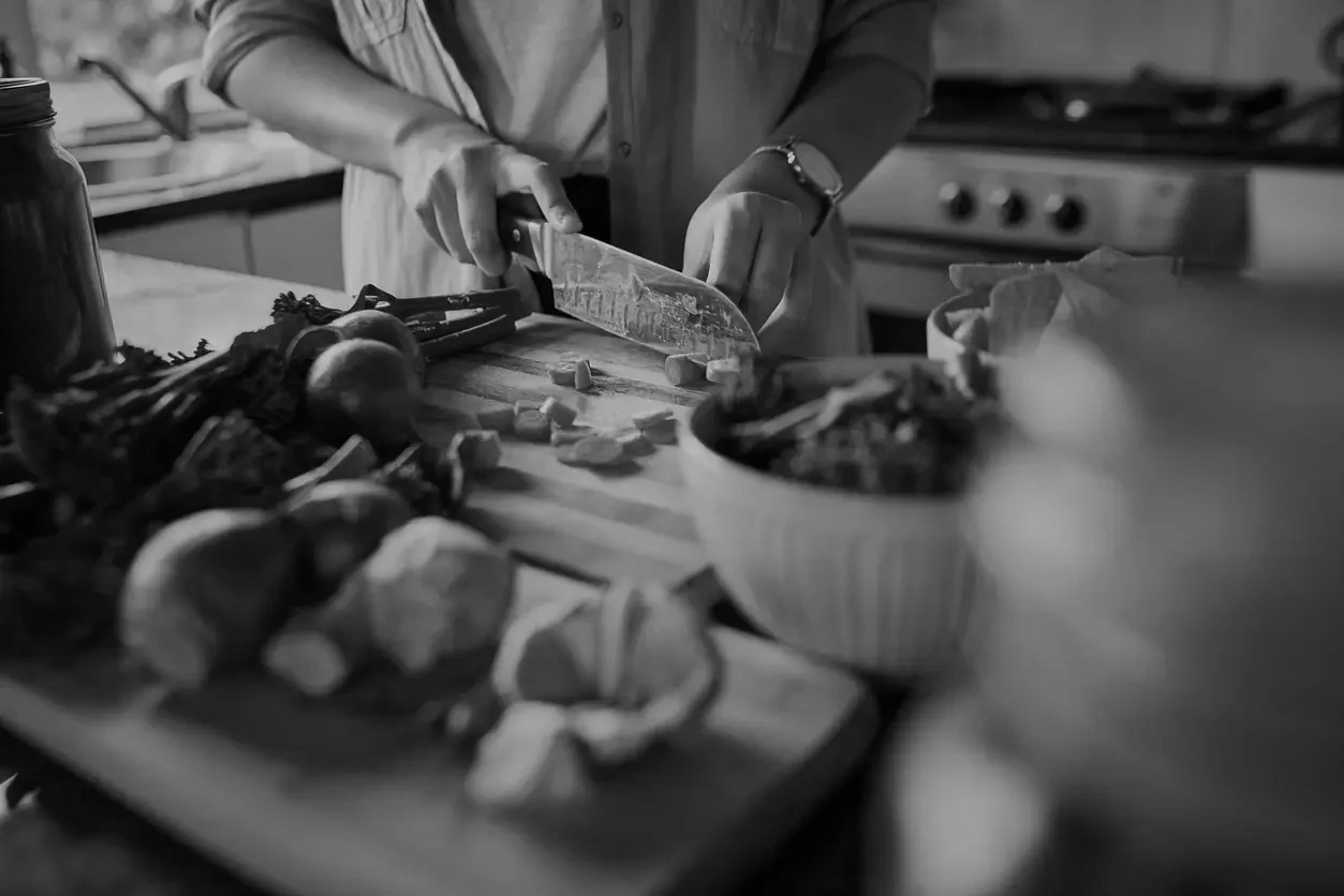 Person chopping vegetables on a wooden cutting board in a kitchen, surrounded by bowls and ingredients, in black and white.