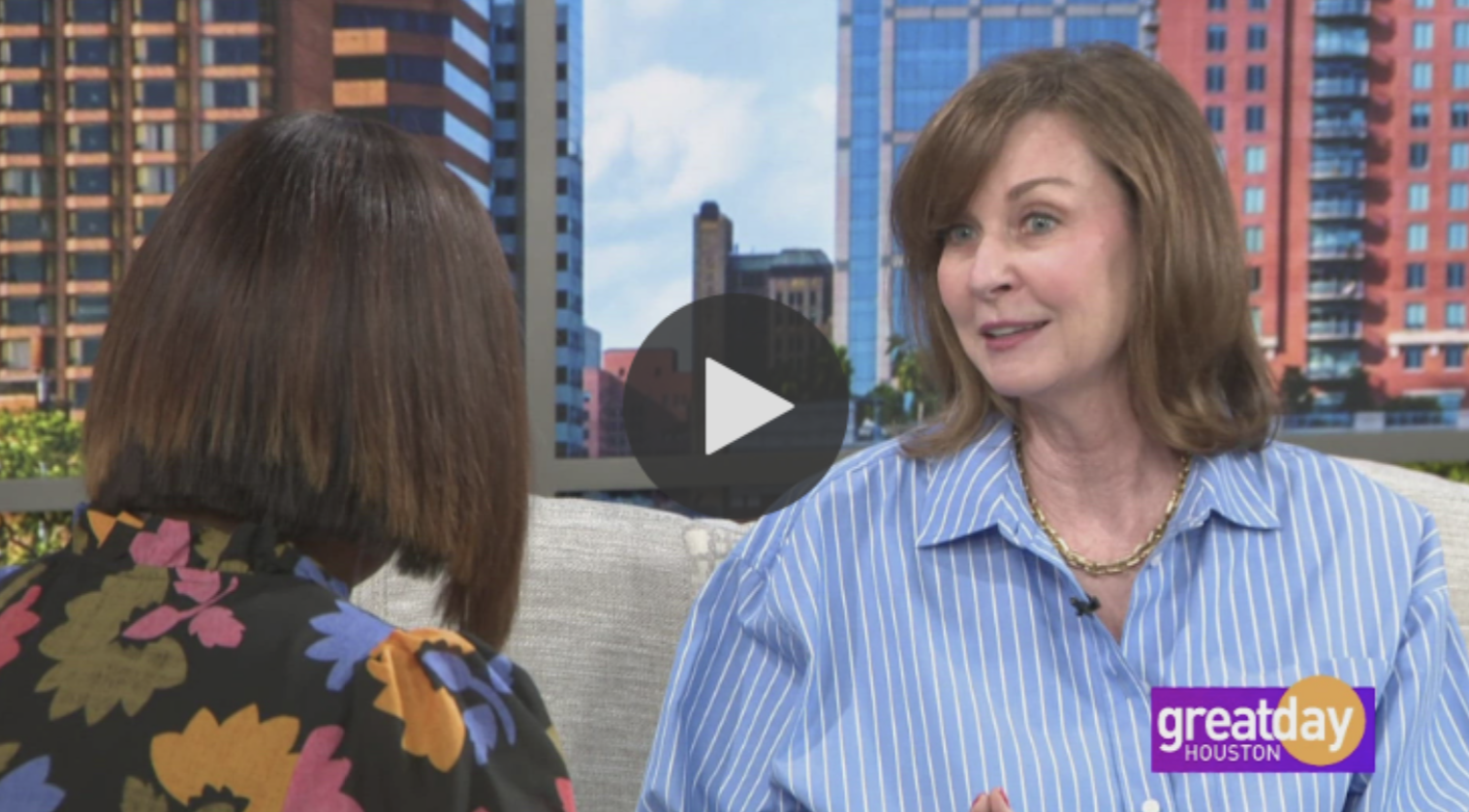 Two women having a conversation on a TV set with a cityscape background. One woman has short brown hair, wearing a black floral top, and the other woman has shoulder-length reddish hair, wearing a blue and white striped shirt with a gold necklace. The logo in the bottom right corner says "Great Day Houston."