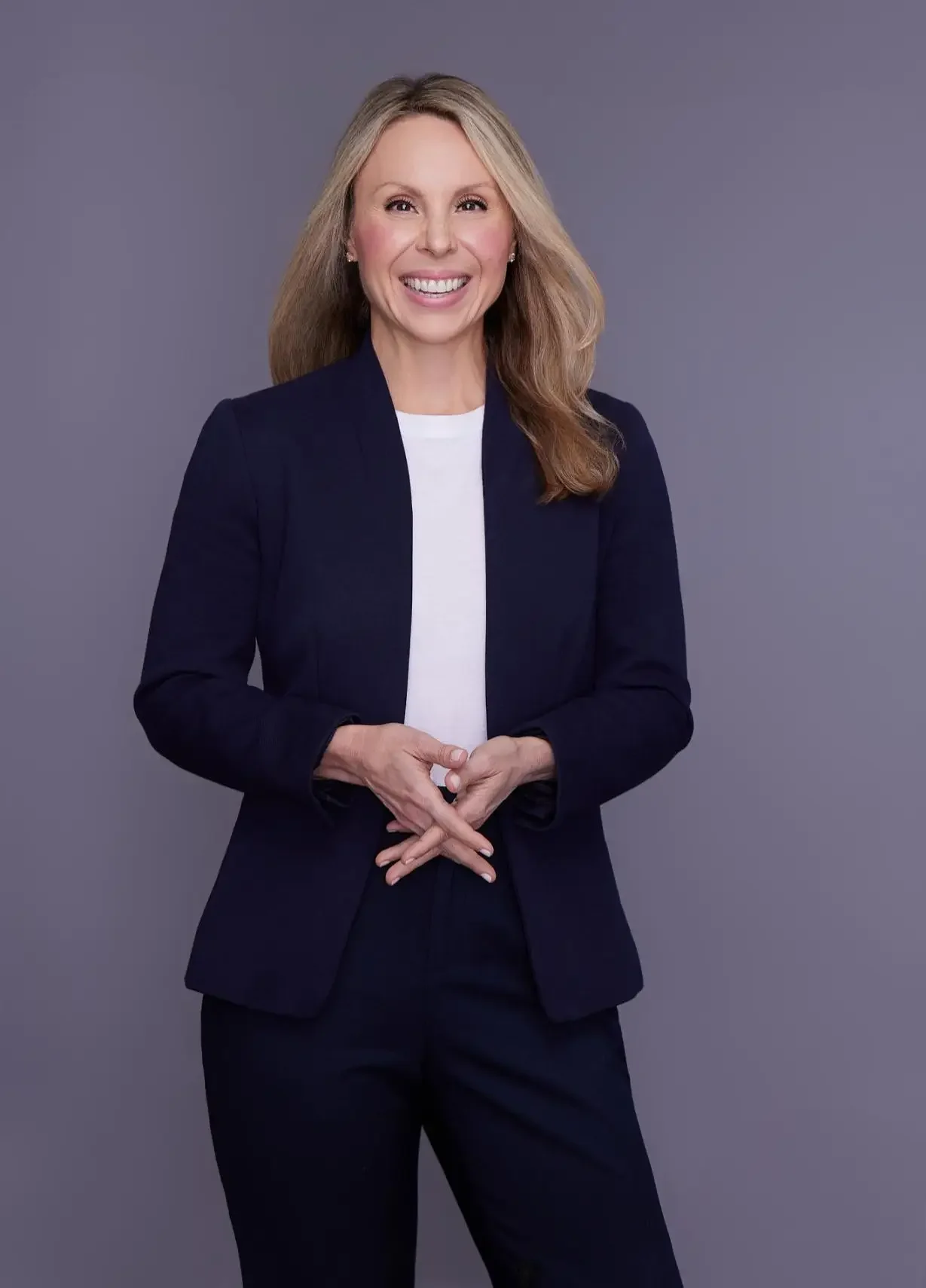 A woman with blonde hair in a navy blazer and white shirt, smiling, standing against a neutral gray background.