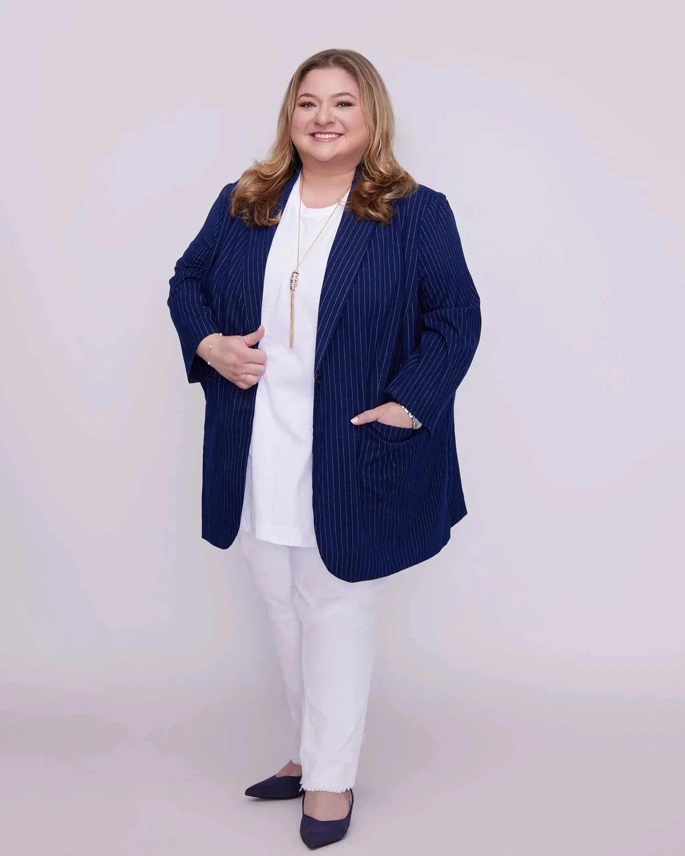 A smiling woman with light brown hair wearing a navy blue pinstripe blazer, white top, white pants, and navy blue heels, standing against a plain light-colored background.