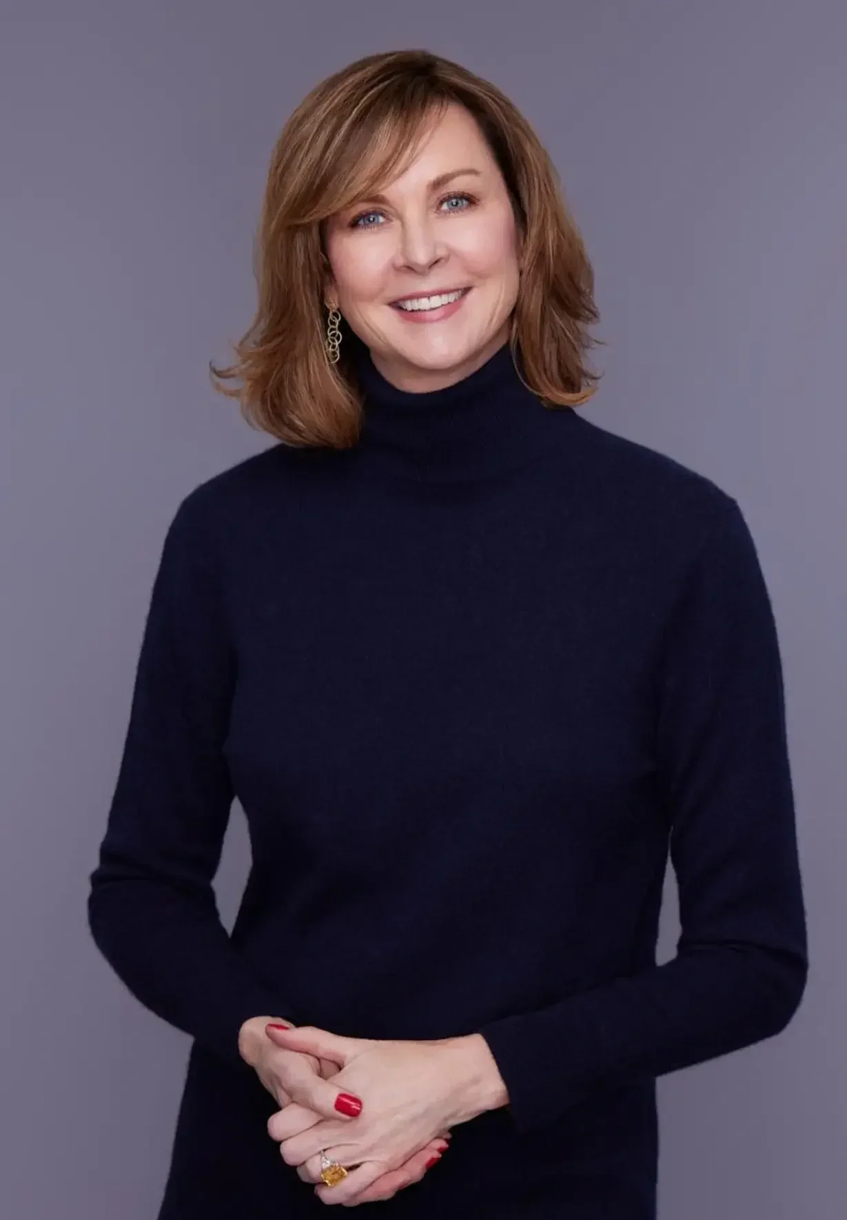 A woman with shoulder-length reddish-brown hair wearing a black turtleneck and earrings, smiling and standing against a gray background.