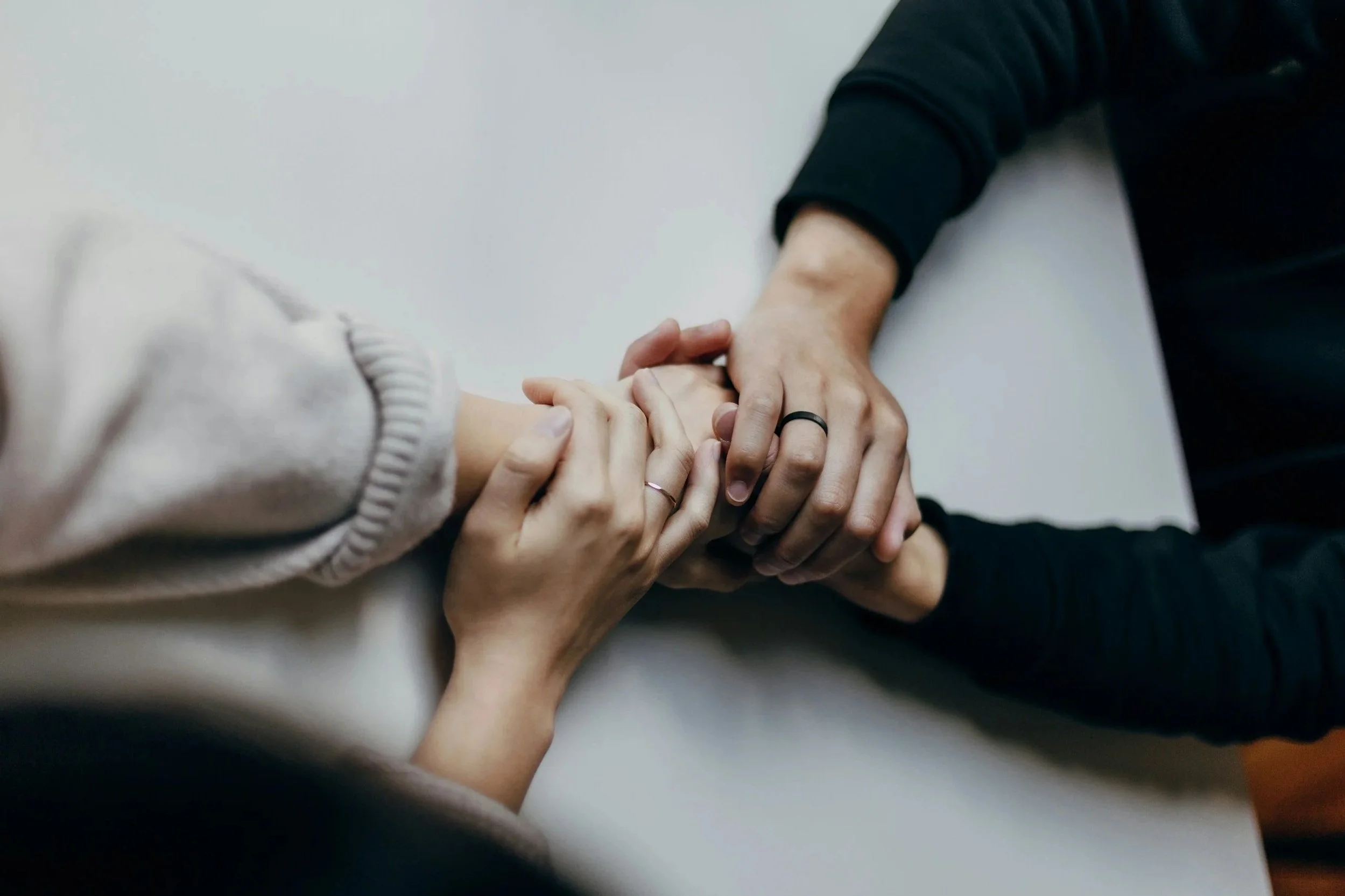 Two people holding hands, one with a lighter skin tone and wearing a beige sweater, the other with a darker skin tone and wearing a black long-sleeve shirt, with rings on their fingers.