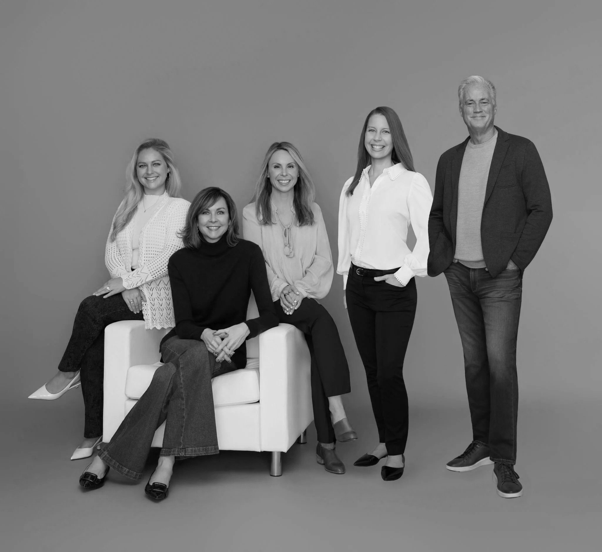 Group of five professionally dressed people, four women and one man, posing together against a plain background. One woman is seated on a white chair, and the others are standing beside her.