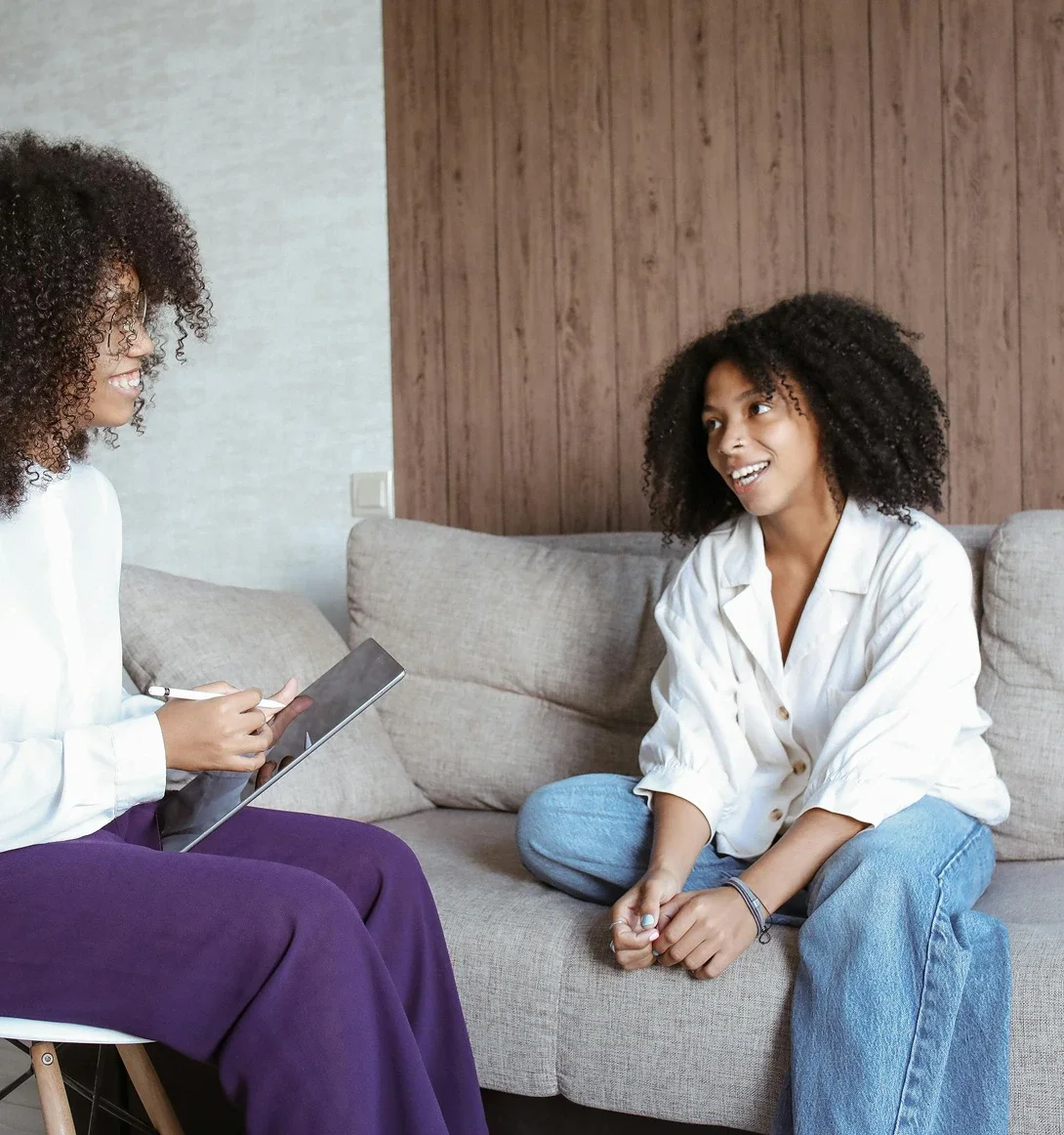 A woman with curly hair and a white shirt smiling and talking to another woman with curly hair, a white shirt, and blue jeans sitting on a gray sofa in a living room.