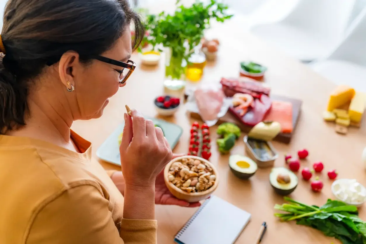 A woman with glasses and earrings holding a bowl of mixed nuts, surrounded by various fresh ingredients such as meats, cheeses, vegetables, and berries on a table.