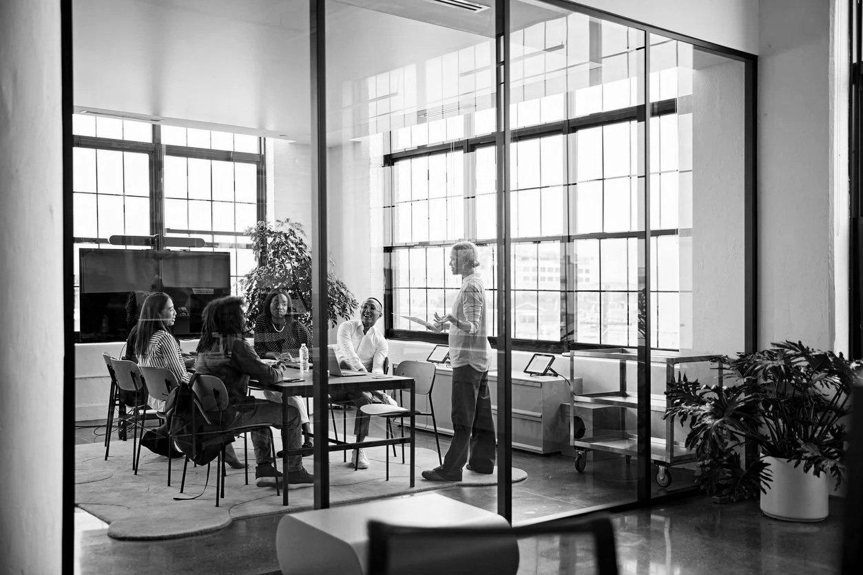 Black and white photo of a business meeting in a modern office conference room with large windows, six women seated at a table, and one woman standing, leading the discussion.