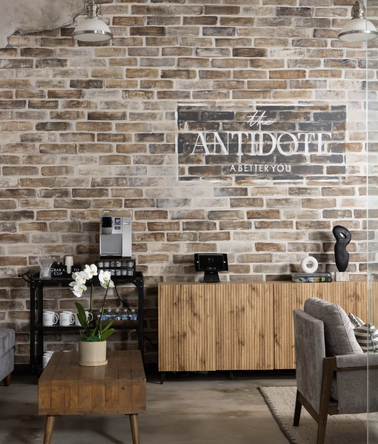 Interior of a lobby with a brick wall, decorative sign, and various furniture and decorations including a potted orchid, coffee machine, and chairs.
