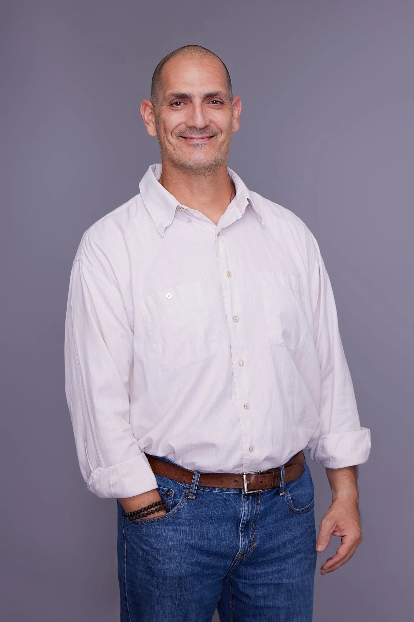 A man with a shaved head and a slight smile wearing a white button-up shirt with rolled sleeves and blue jeans, standing against a gray background