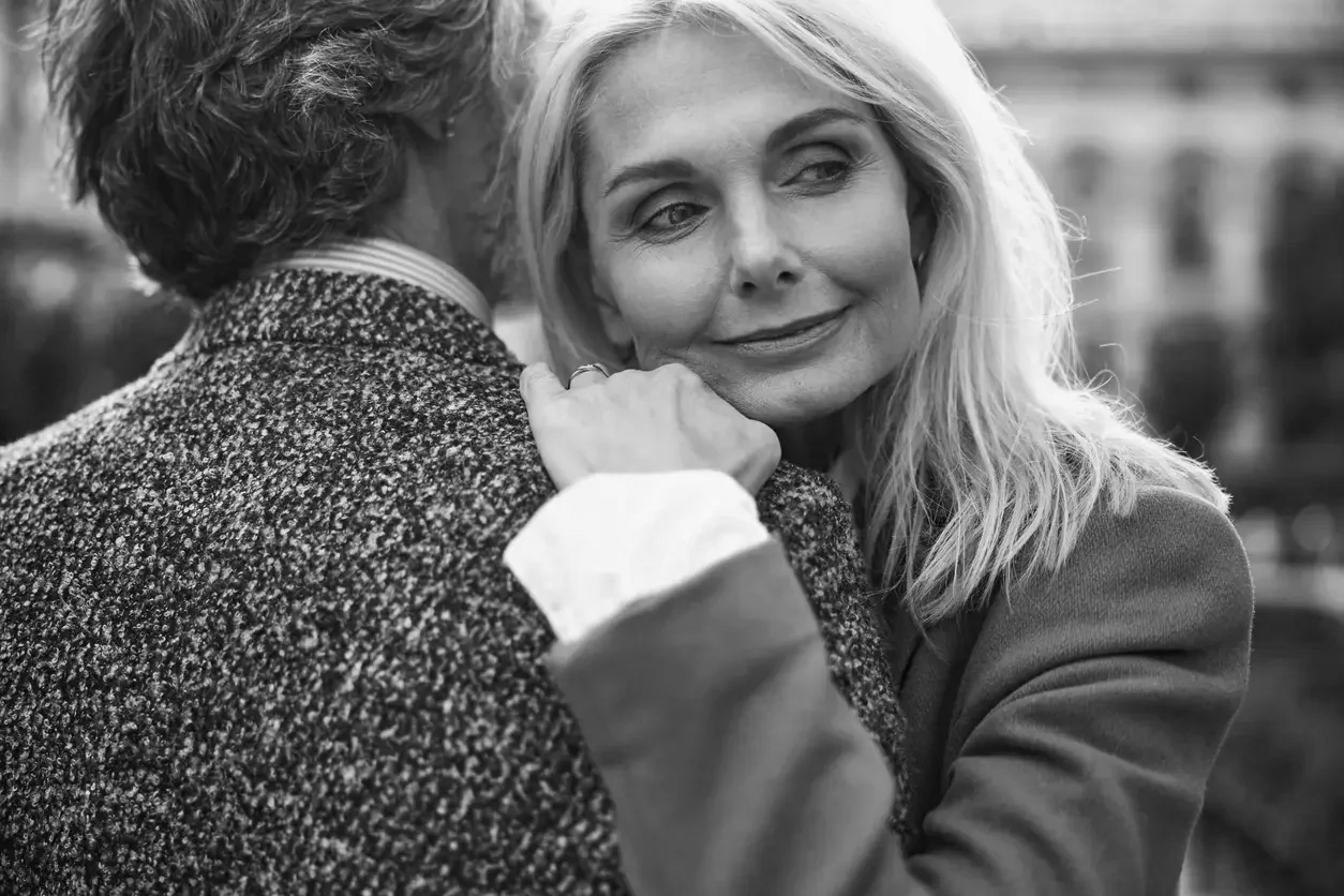 A woman with blonde hair smiles softly as she gently touches a man's shoulder, whose face is turned away from the camera, in a close embrace.