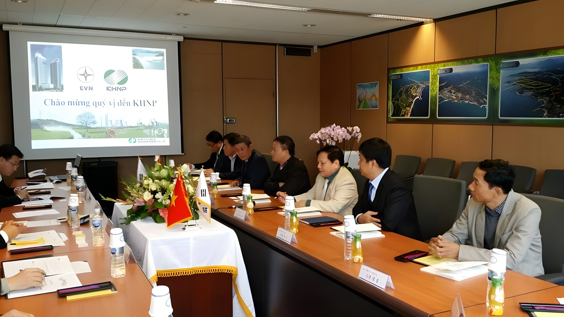 A formal meeting room with several people sitting around a large wooden conference table. There is a projector screen at the front displaying a presentation with logos and Vietnamese text. The table is decorated with a floral arrangement, flags of Vi