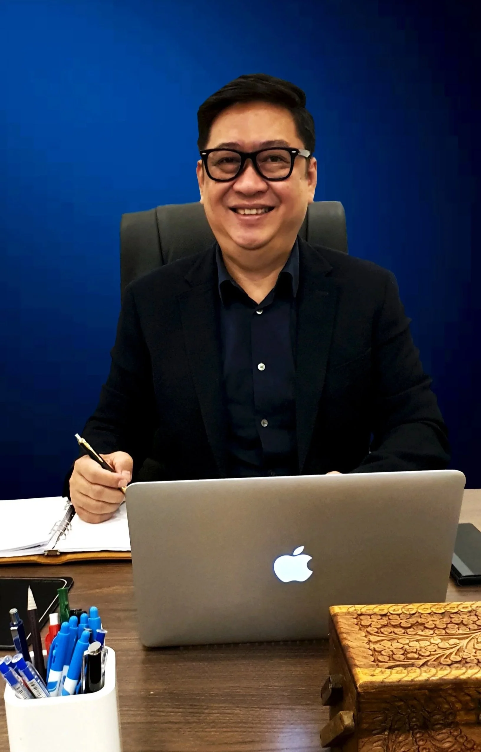 Man wearing black suit and glasses sitting at desk with MacBook, smiling, with blue wall background.