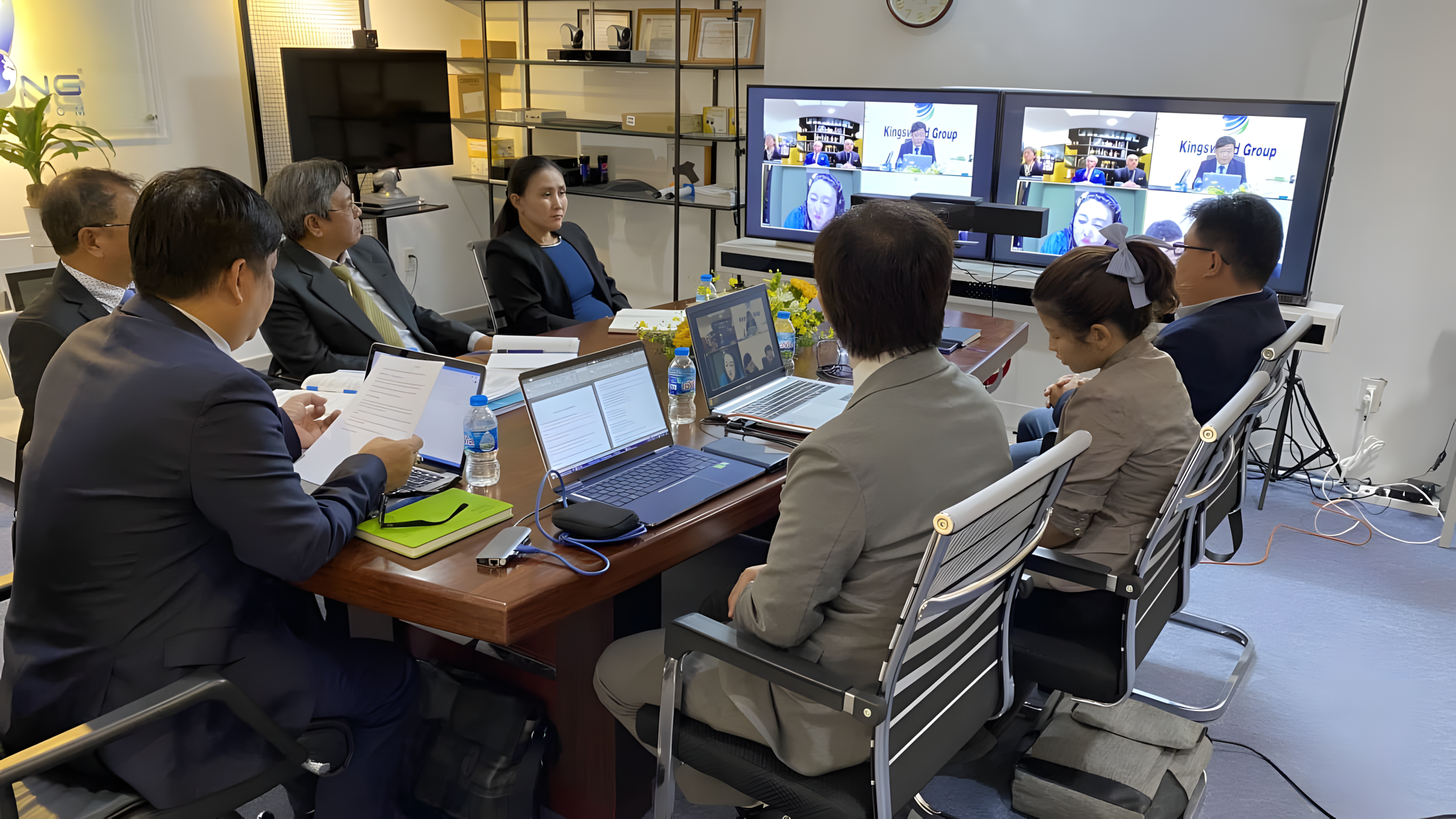 Business meeting in a conference room with nine people seated around a wooden table, working on laptops and reviewing documents, while participating in a video conference displayed on a large screen at the front of the room.