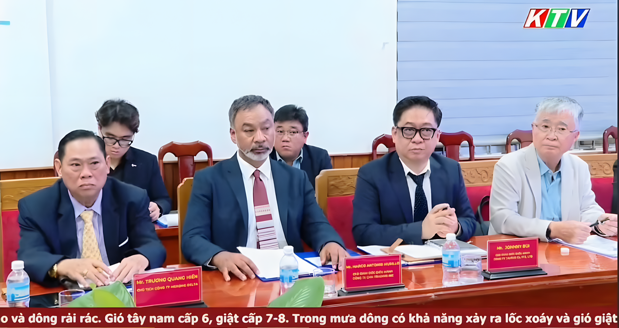 A group of six men and one woman sitting at a conference table during a meeting. They are dressed in formal business attire. There are nameplates, documents, water bottles, and writing materials on the table. In the background, there is a whiteboard and a displayed logo in the upper right corner.