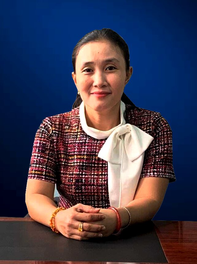 A woman sitting at a table with a dark tablecloth, wearing a patterned dress with a white bow tie around her collar, and bracelets on her wrists, against a plain dark blue background.