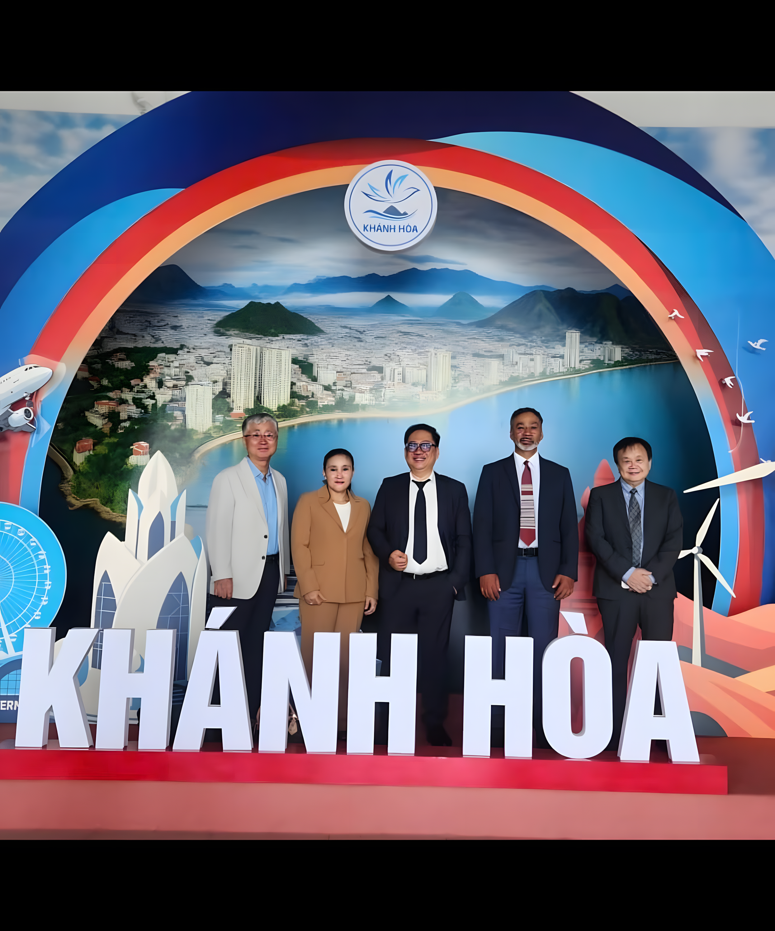 A group of five people standing in front of a backdrop featuring a cityscape and landscape, with large letters spelling 'Khánh Hòa' in front of them.