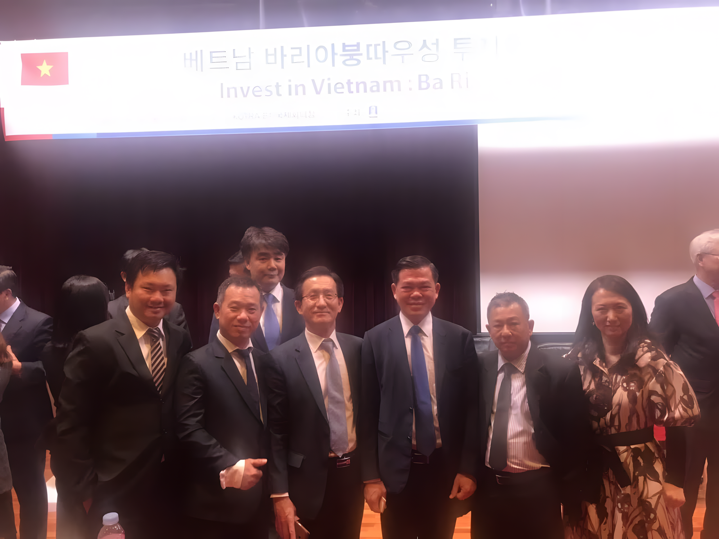 Group of eight people in business attire standing in front of a large screen with a Vietnam flag and text about investment in Vietnam.