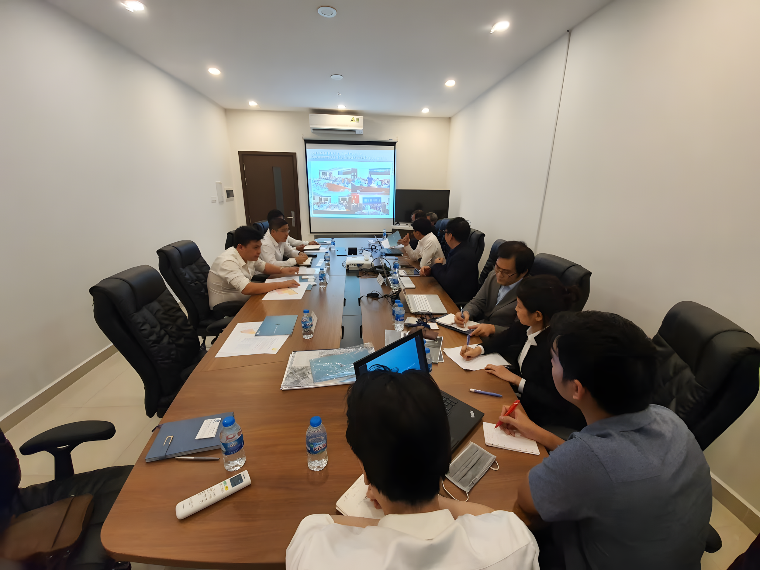 A group of professionals sitting around a conference table in a meeting room, watching a presentation on a large screen.