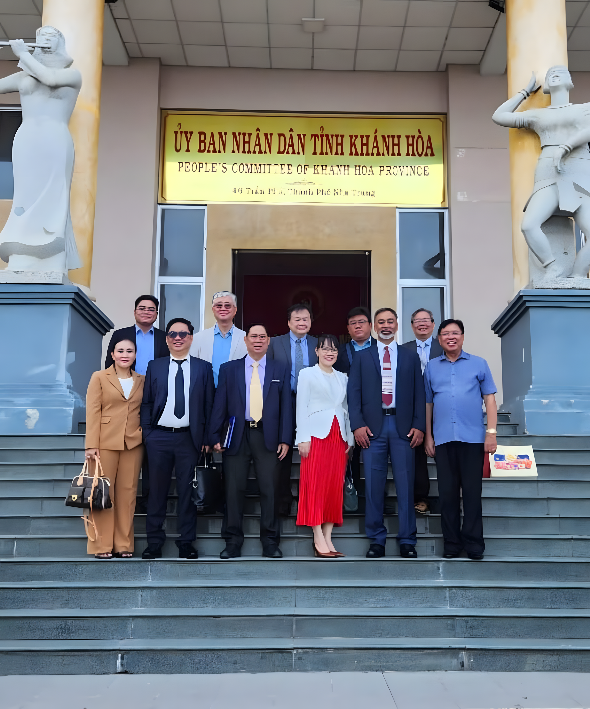 Group of ten people in formal attire standing on steps outside the People's Committee of Khanh Hoa Province building, with statues on either side and a yellow sign overhead.