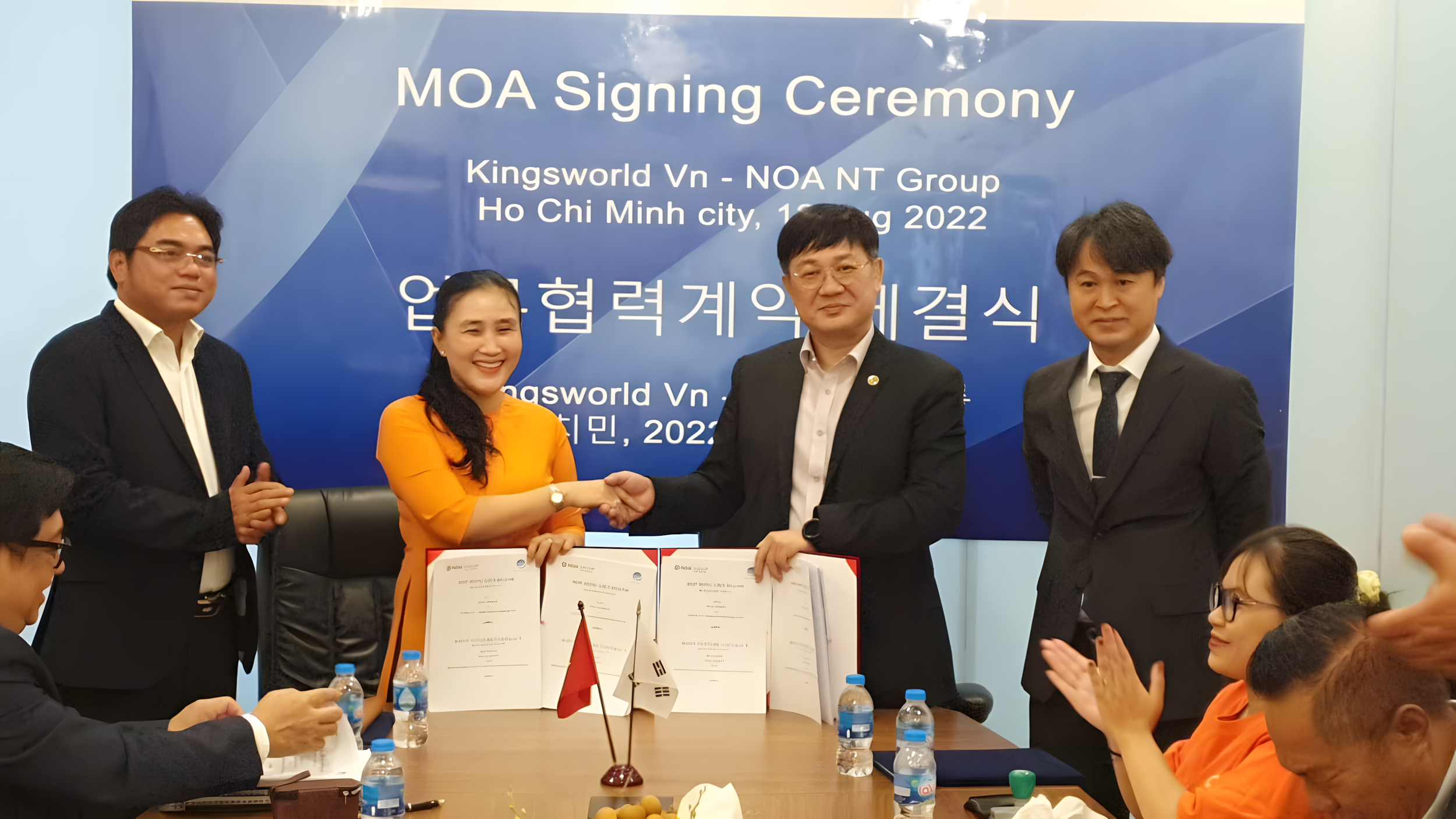 Two people shake hands at a signing ceremony, flanked by two men and several seated attendees, with a blue backdrop displaying event details.