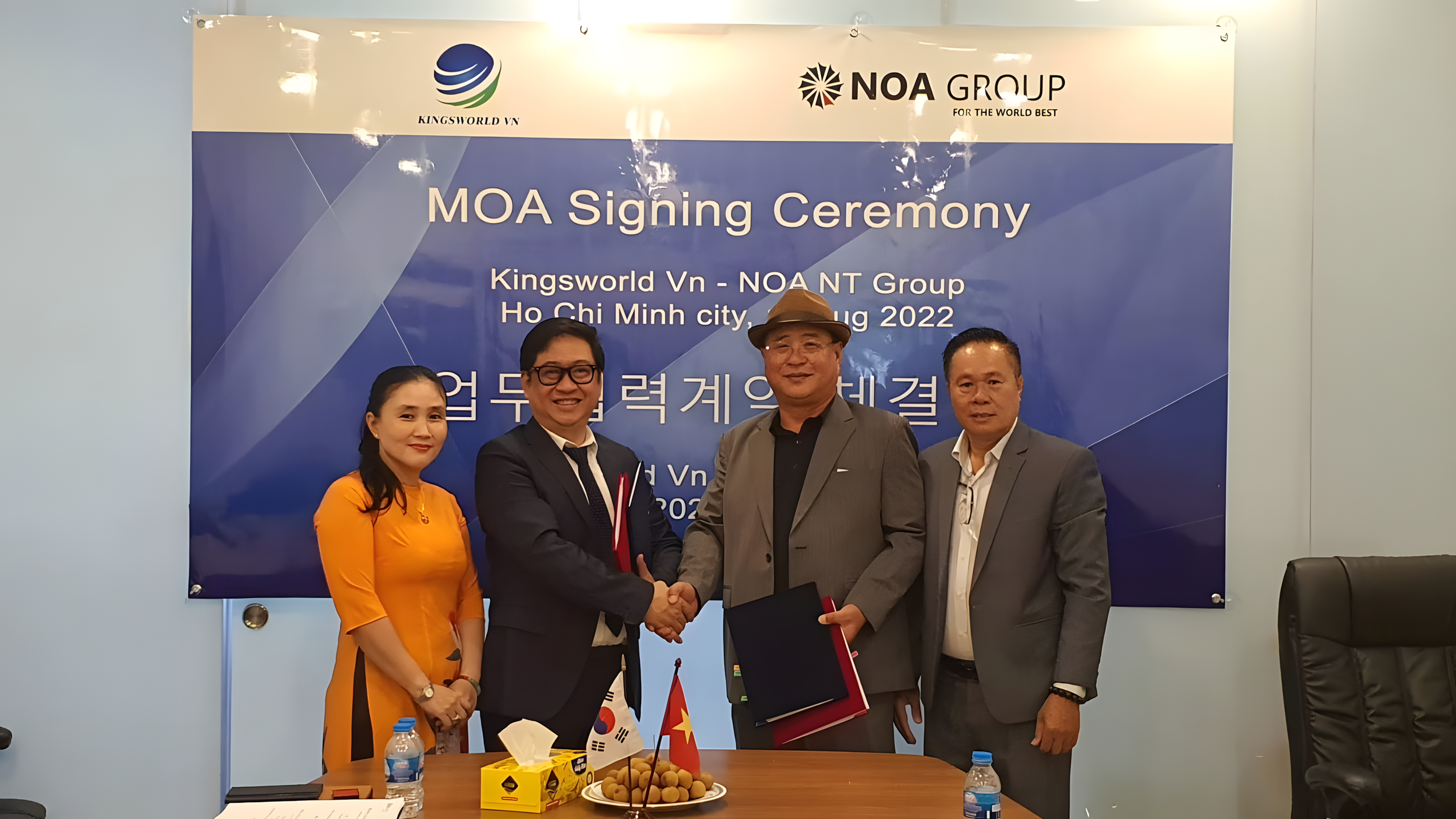 Four people standing in front of a sign at a signing ceremony, two of whom are shaking hands. The sign reads 'MOA Signing Ceremony' and 'Kingsworld Vn - NOA NT Group, Ho Chi Minh City, August 2022.' There are two small flags on the table: South Korea