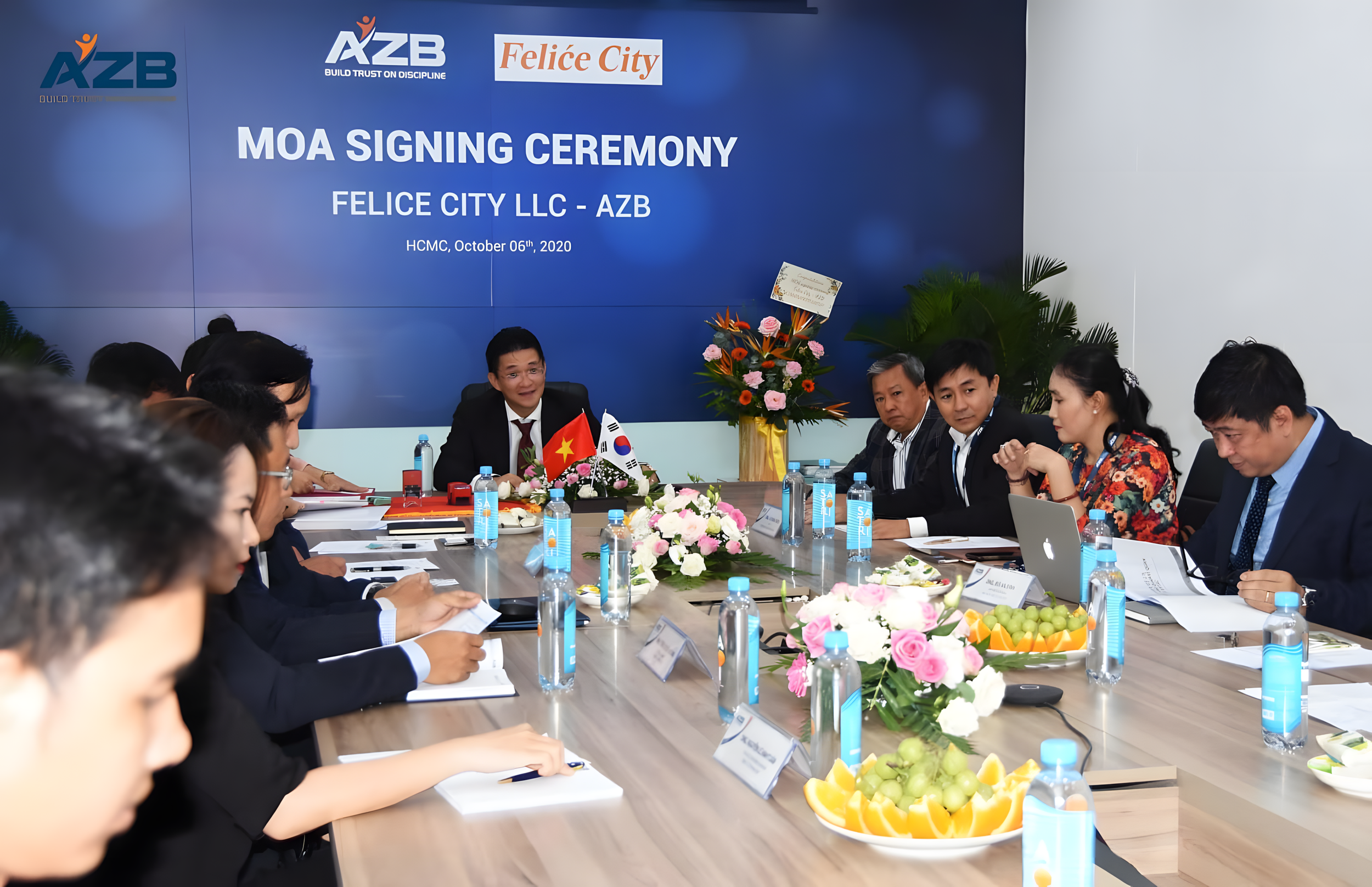 Meeting room with people attending Felice City LLC - AZB MOA signing ceremony, seated around a table with flowers, bottled water, laptops, and documents, with a backdrop displaying the event details and the logos of AZB and Felice City.