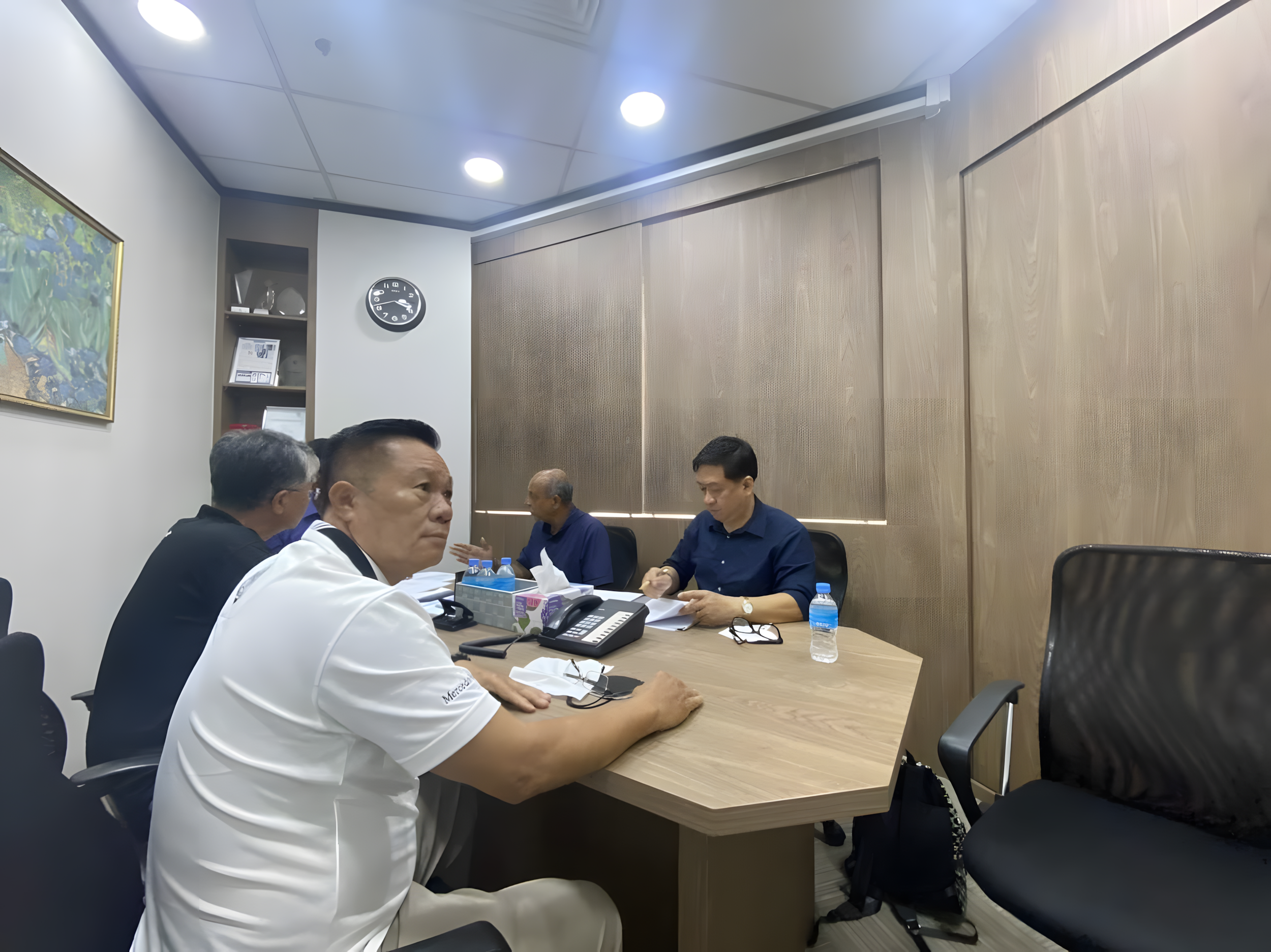 Meeting room with four men seated at a conference table, one in a white shirt, others in dark blue shirts, with water bottles, tissues, and documents on the table.